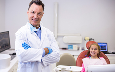 A dentist smiling with a young girl sitting in a dental chair in the background
